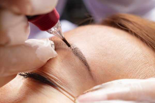 Beauty master performs permanent eyebrow makeup in a beauty salon, close-up. Hands of a cosmetologist doing microblading of eyebrows. Tattoo on the face.