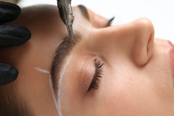 Beautician making permanent eyebrow makeup to young woman on white background, closeup